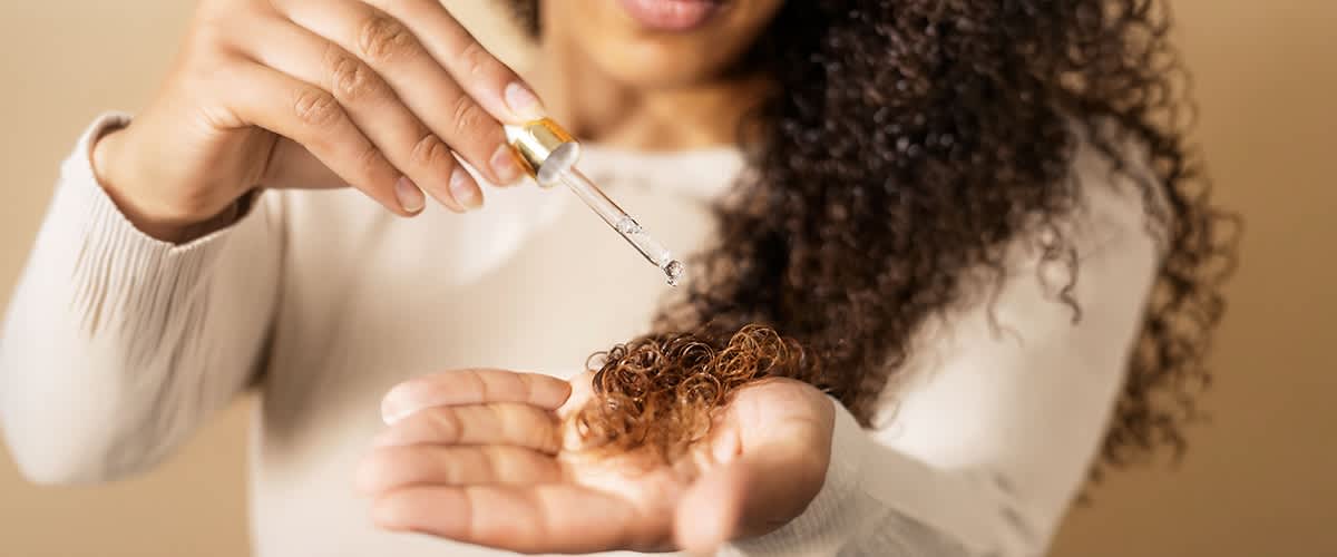 Hand gibt einige Lockensträhnen in die andere Hand, darüber eine Pipette mit klarem Öl, Tropfen fällt auf die Haare