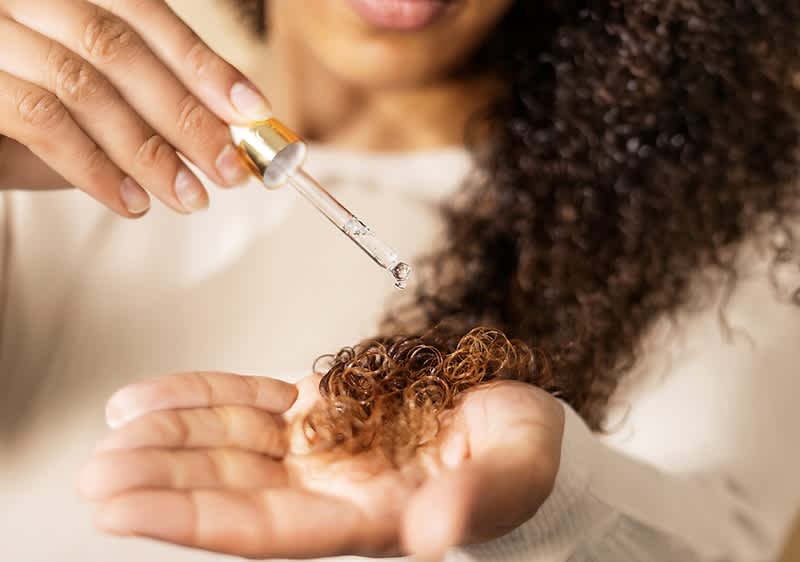 Hand mit Lockensträhne hält Haaröltröpfchen aus einer Pipette über der offenen Handfläche, im Hintergrund lächelnde Person mit dichtem lockigem Haar