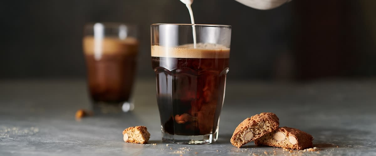 Ein Glas schwarzer Kaffee mit Crema, in das Milch gegossen wird, vorne rechts drei Mandelkekse, links ein Keksstück auf grauem Untergrund, im Hintergrund unscharfes zweites Glas mit Kaffee