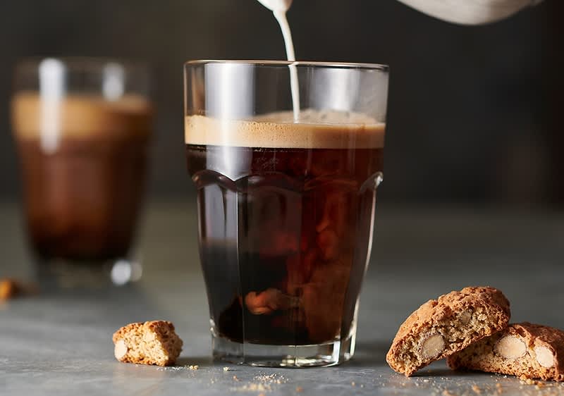 Ein Glas schwarzer Kaffee mit feiner Crema, in das oben Milch gegossen wird, im Vordergrund zwei Mandelbiscotti und ein zerbrochenes Stück auf grauem Untergrund, im Hintergrund unscharfes zweites Glas Kaffee