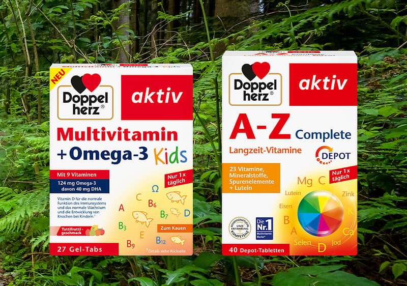 Zwei Doppelherz aktiv Produktpackungen nebeneinander auf Waldboden: links weiße Box „Multivitamin + Omega-3 Kids“ mit bunten Schriftzügen und 27 Gel-Tabs, rechts weiße Box „A–Z Complete Langzeit-Vitamine“ mit Farbrad und 40 Depot-Tabletten