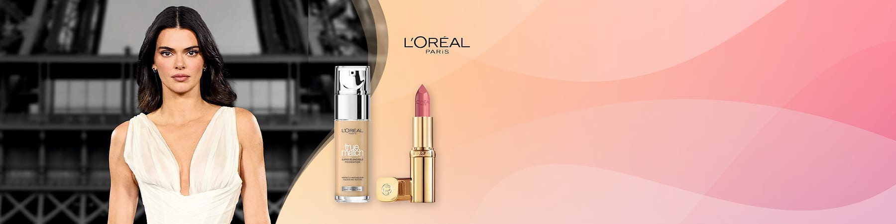 Eine Frau mit dunklem kinnlangen Haar und weißem ärmellosen Kleid steht links, rechts daneben eine Glasflasche L'Oréal True Match Foundation mit silberner Pumpe und eine goldene L'Oréal-Lippenstift-Hülse mit rosa Lippenstift vor rosa-beigem Hintergrund, oben rechts das L'Oréal Paris-Logo