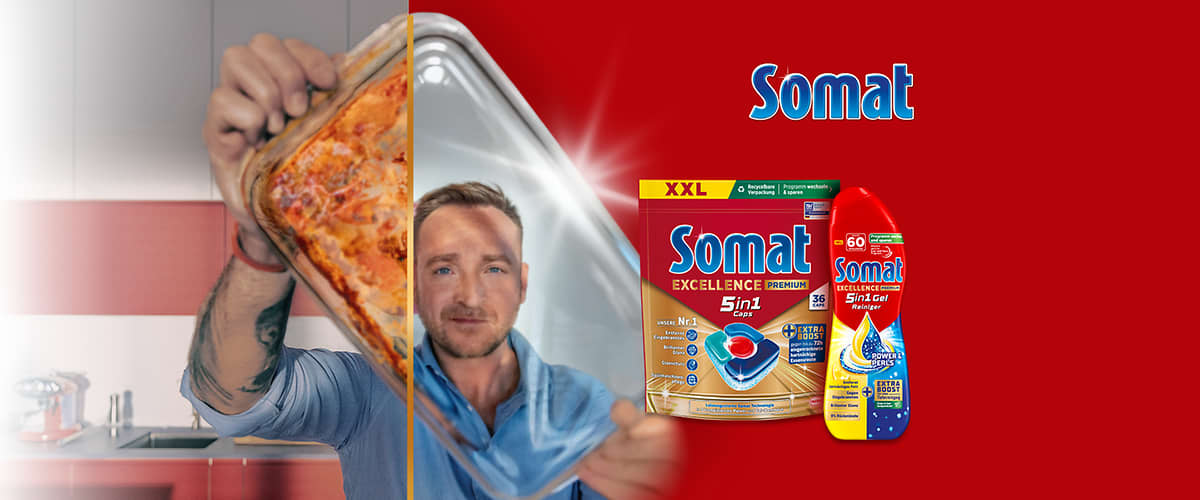 Werbewand mit rotem Hintergrund, links eine Hand hält eine Auflaufform mit gebackenem Gericht, daneben ein Mann in blauem Hemd zieht transparenten Frischhaltefilm ab, rechts zwei Somat-Produktverpackungen: eine XXL-Packung Somat Excellence 5in1 Caps und eine rote Somat Spülmaschinen-Gel-Flasche, oben rechts das Somat-Logo