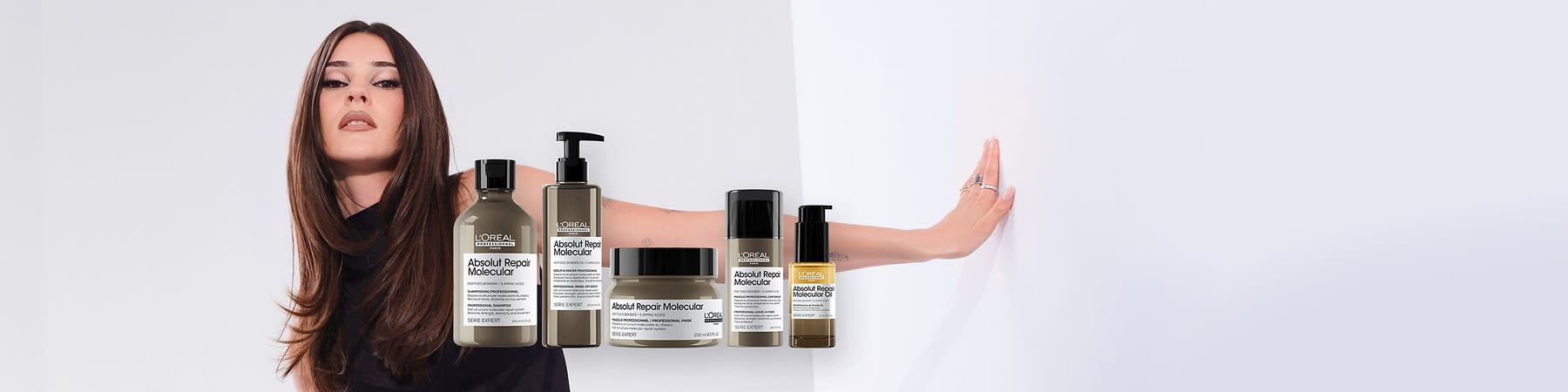 Eine junge Frau mit langen braunen Haaren und schwarzem Oberteil streckt einen Arm zur Seite, davor fünf L’Oréal Absolut Repair Molecular Haarpflegeprodukte in verschiedenen Pumpspendern und Tiegeln mit sichtbaren Produktetiketten