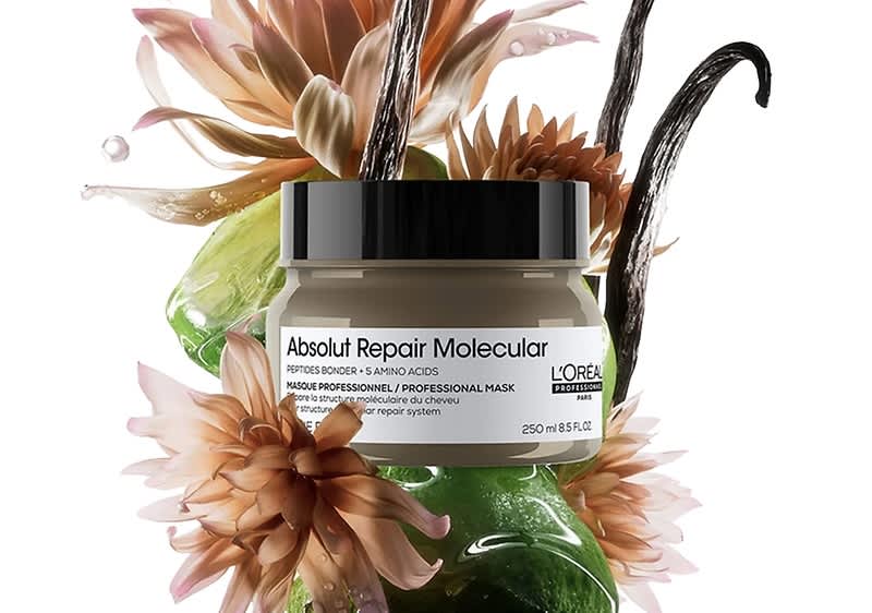 Ein runder Tiegel mit schwarzem Deckel und weißem Etikett mit der Aufschrift „Absolut Repair Molecular“ von L’Oréal, platziert vor grünen Gelkissen, Vanilleschoten und beige-pinken Blüten