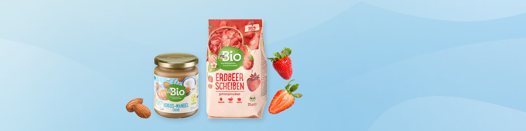 Links ein Glas dmBio Kokos-Mandel-Creme mit goldfarbenem Deckel und sichtbarem Etikett, rechts eine Packung dmBio Erdbeer Scheiben gefriergetrocknet 25 g mit rotem Design und abgebildeten Erdbeeren, Mandeln und aufgeschnittene Erdbeere