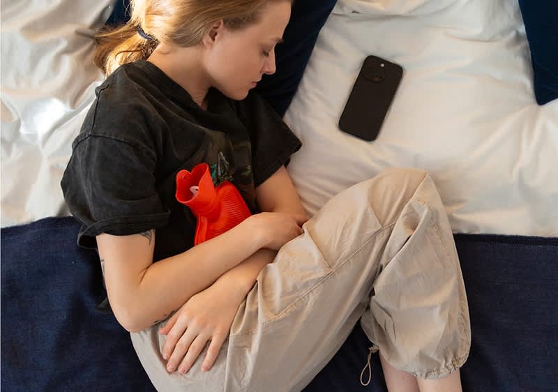 Junge Person liegt seitlich auf einem Bett und hält eine rote Wärmflasche an den Bauch, ein schwarzes Smartphone liegt neben dem Kopf auf dem Kissen