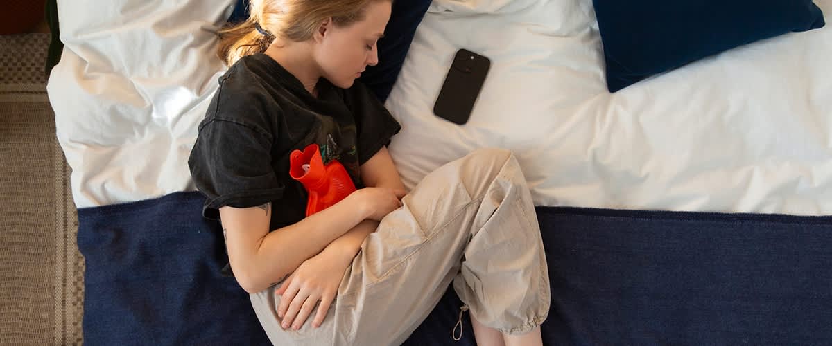 Junge Person liegt seitlich auf einem Bett und hält eine rote Wärmflasche an den Bauch, ein schwarzes Smartphone liegt neben dem Kopf auf dem Kissen