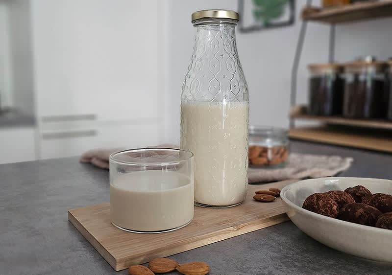 Glasflasche mit hellem Pflanzenmilch-Getränk und ein niedriges Glas mit der selben Milch auf einem Holzbrett, daneben einige ganze Mandeln und eine Schüssel mit Schokotrüffeln in einer Küche