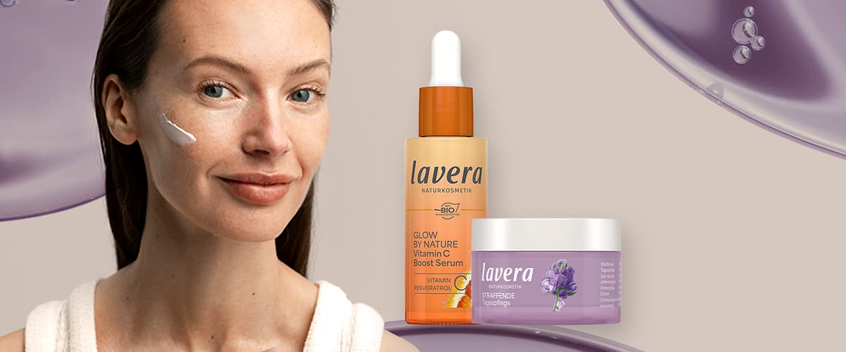 Porträt einer lächelnden Frau mit aufgetragener Gesichtscreme am rechten Wangenknochen, daneben eine orangefarbene Lavera-Serumflasche mit Pipette und Beschriftung „Glow by Nature Vitamin C Boost Serum“ sowie ein lila-weißes Lavera-Gesichtspflegeglas mit Blütenillustration und Marke Lavera
