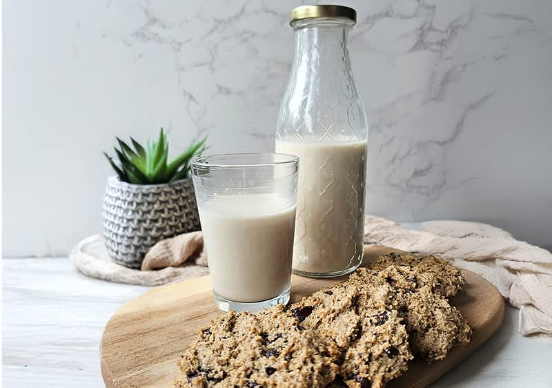 Glasflasche mit heller Pflanzendrink und ein gefülltes Trinkglas auf einem Holzbrett, daneben mehrere Haferkekse mit Schokostückchen, im Hintergrund ein Topf mit kleiner Sukkulente und ein beige Leinenstoff