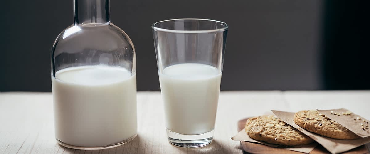 Ein Glas Milch zur Hälfte gefüllt, links eine bauchige Glasflasche mit Milch und rechts auf einem Holzbrett zwei Haferkekse auf Backpapier ohne Punkt am Satzende