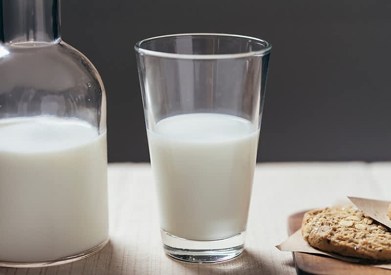 Glas mit Milch zur Hälfte gefüllt in der Bildmitte, links eine Glaskaraffe mit Milch und rechts auf einem Holzbrett Haferkekse und ein Messer auf einer hellen Tischoberfläche
