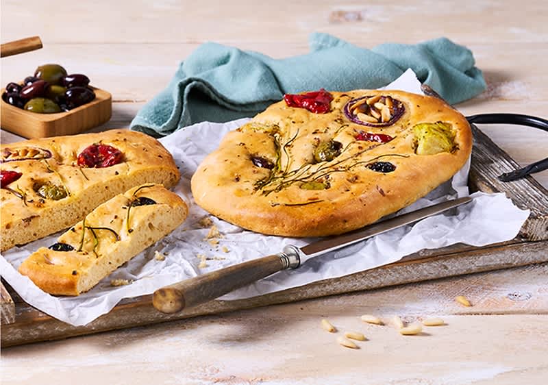 Runde, goldbraune Focaccia mit Rosmarin, schwarzen Oliven, halbierten Cocktailtomaten und roten Zwiebelringen auf Backpapier und Holzbrett, daneben ein Messer und eine Schale gemischter Oliven