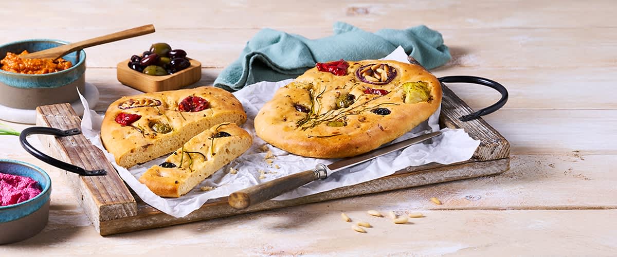 Runde, goldbraune Focaccia mit Rosmarin, schwarzen Oliven, halbierten Cocktailtomaten und roten Zwiebelringen auf Backpapier und Holzbrett, daneben ein Messer und eine Schale gemischter Oliven