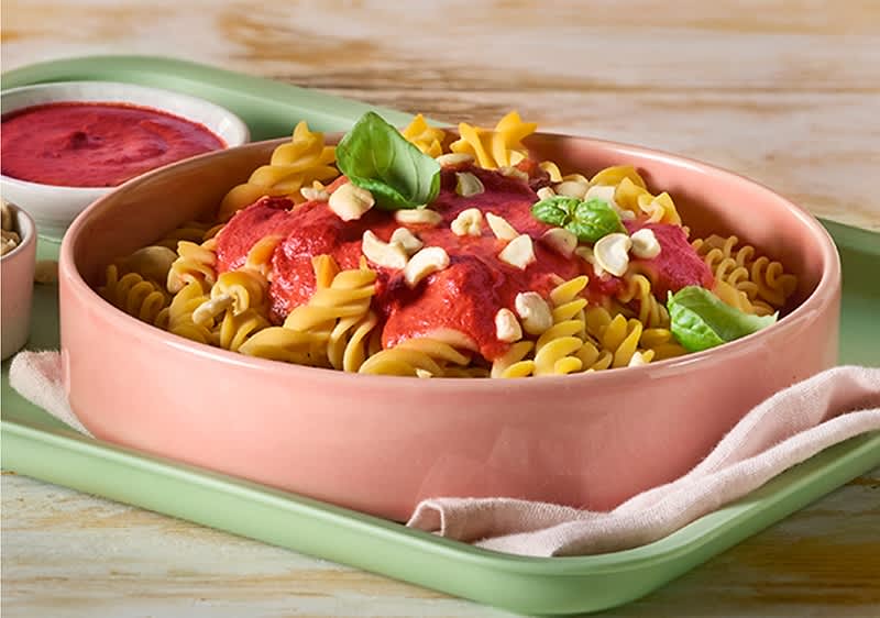 Gekochte Fusilli-Nudeln in einer flachen rosa Schale mit roter Tomatensauce, gehackten Nüssen und frischen Basilikumblättern auf einem hellgrünen Tablett ohne Punkt