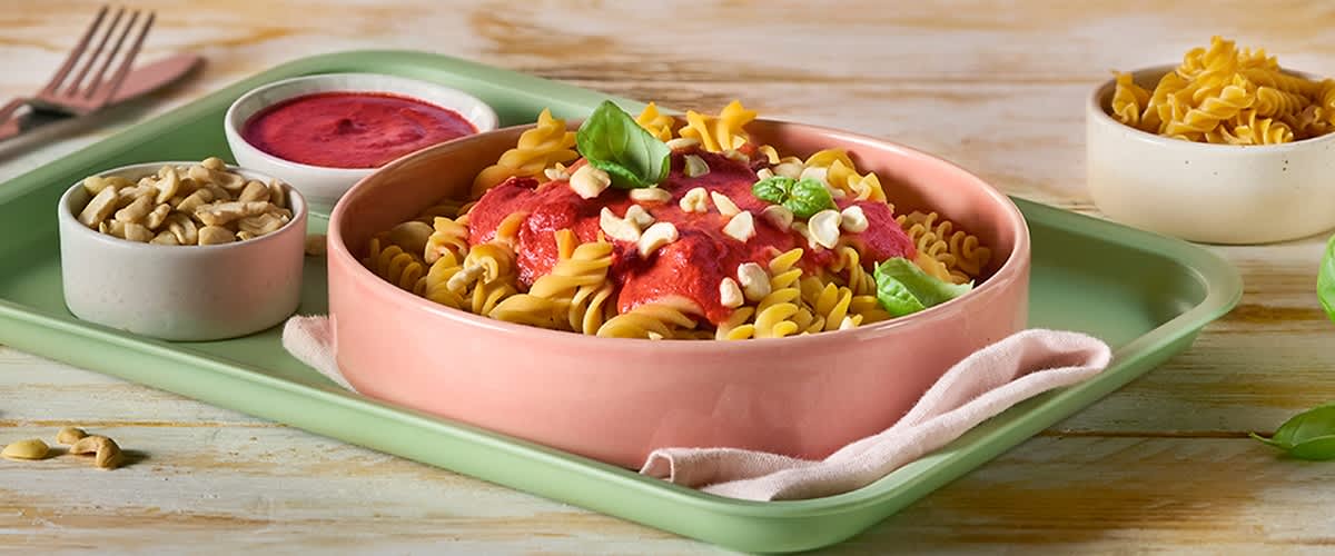 Gekochte Fusilli-Nudeln in einer flachen rosa Schale mit roter Tomatensauce, gehackten Nüssen und frischen Basilikumblättern auf einem hellgrünen Tablett ohne Punkt