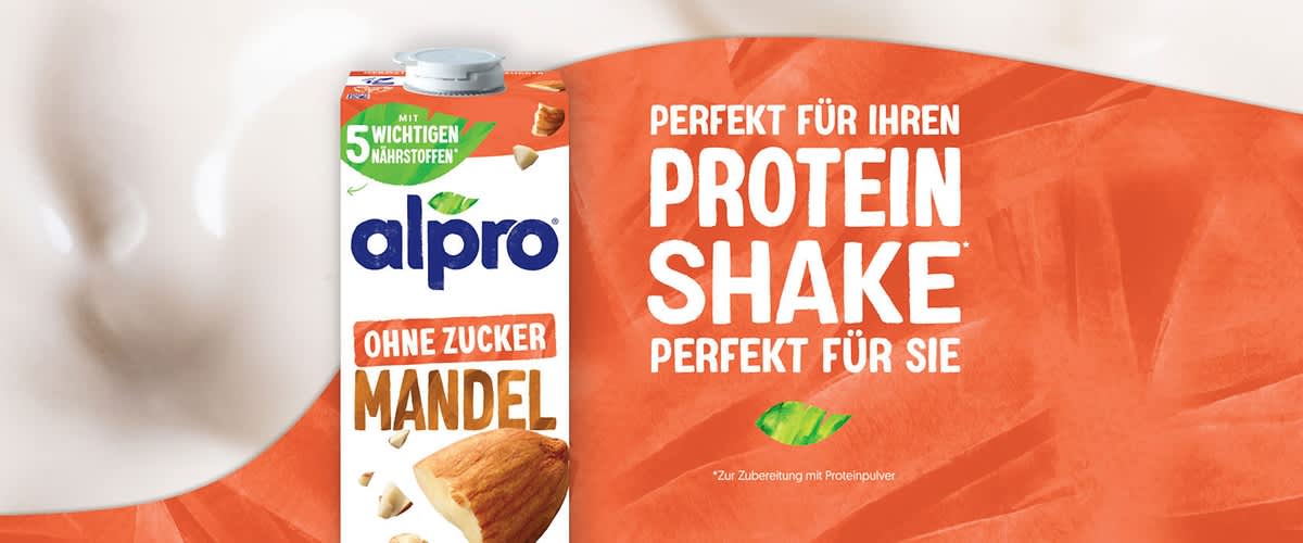 Alpro No Sugars Nutty Almond Getränkepackung neben einer Auflaufform mit gebackenen Blaubeer-Haferstücken, verstreuten Mandeln und einer kleinen weißen Milchkännchen im Hintergrund