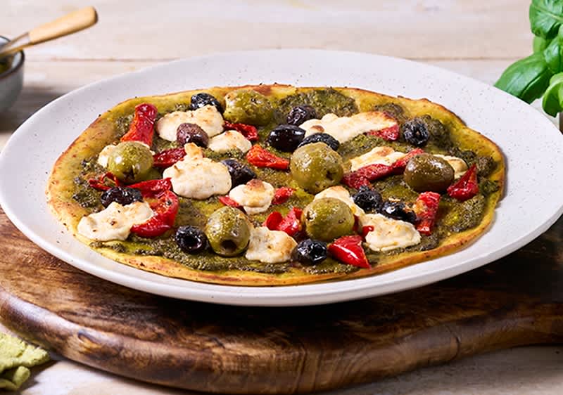 Flache Pesto-Pizza auf weißem Teller mit grünen und schwarzen Oliven, getrockneten Tomaten und kleinen Mozzarellakugeln auf Holzunterlage