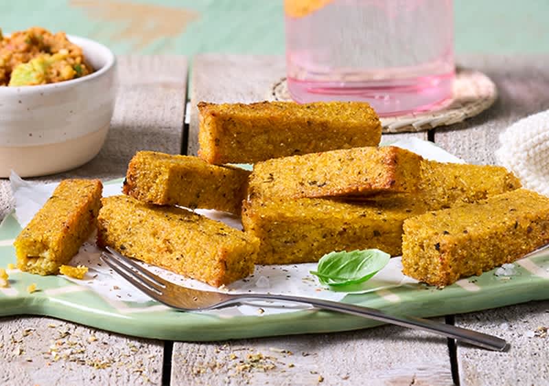 Mehrere goldbraune Polenta-Stangen auf weißem Backpapier auf einem hellgrünen Teller, daneben eine Gabel und ein Basilikumblatt, im Hintergrund eine Schale Dipp und ein Glas mit rosa Flüssigkeit