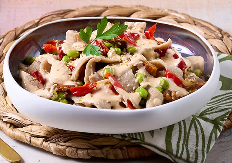 Weiße Schale mit dünn geschnittenem Fleisch, grünen Erbsen und roten Paprikastreifen, überzogen mit cremiger heller Sauce und garniert mit Petersilienblättern, auf geflochtenem Untersetzer und Stoffserviette
