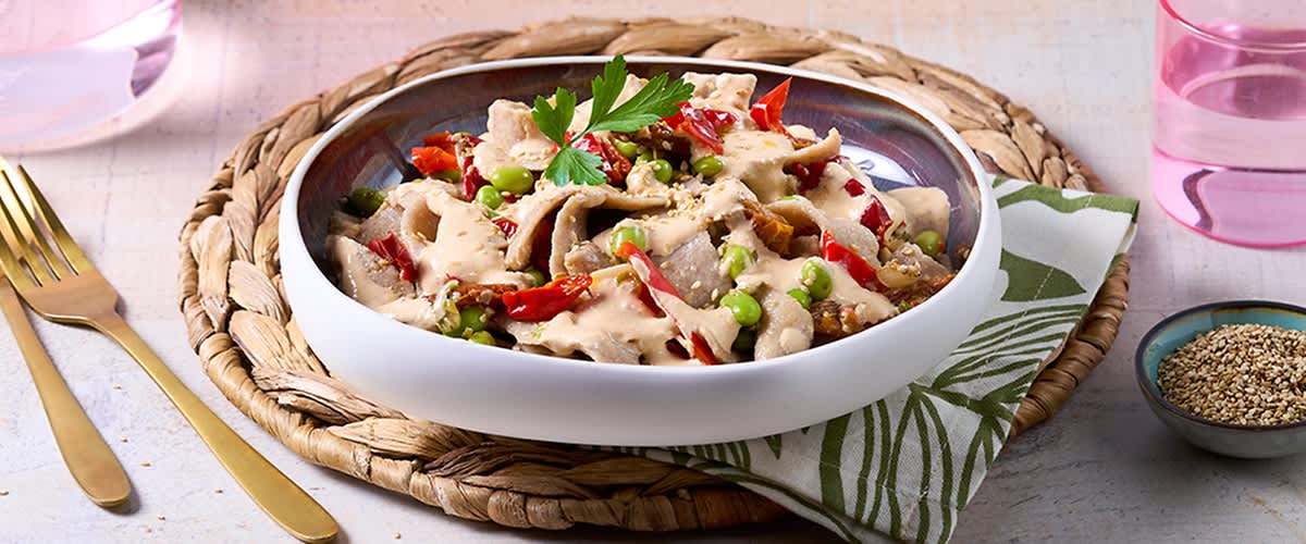 Vollkornnudeln mit edamame, roten Paprikastreifen und cremiger heller Sauce, garniert mit Petersilienblatt in weißer Schale auf geflochtenem Untersetzer und grün-weiß gemustertem Stoffserviette