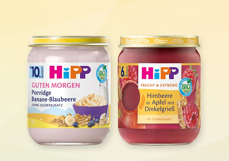 Zwei Gläschen Babynahrung der Marke HiPP nebeneinander: links helles Glas mit Aufschrift „HiPP Guten Morgen Porridge Banane-Blaubeere 10. Monat ohne Zuckerzusatz“ und Bild von Hafer, Banane und Blaubeeren, rechts rotes Glas mit Aufschrift „HiPP Frucht & Getreide Himbeere in Apfel mit Dinkelgrieß 6+ Monate 0% Zuckerzusatz“ und Bild von Himbeeren und Apfel