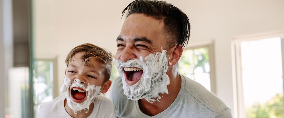 Männerpflege: Mann und Kind mit Rasierschaum lachend vor Spiegel