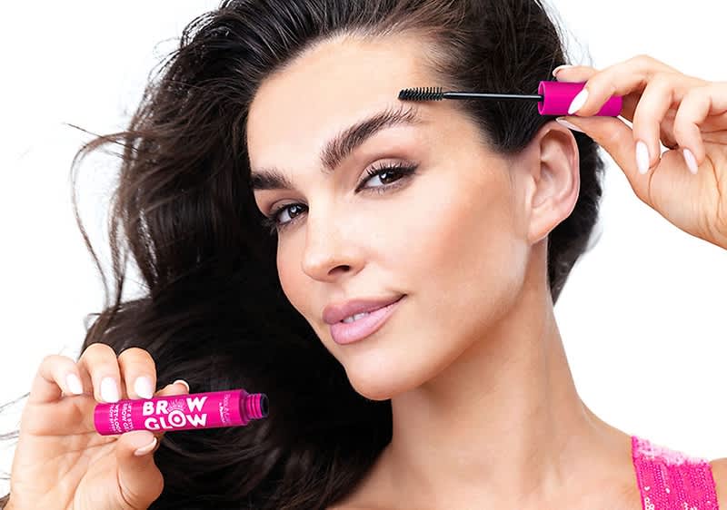Frau hält mit erhobenen Händen zwei pinke Stift-Applikatoren, Beschriftung auf einem: „BROW GLOW“, eine Hand führt Applikator zur Augenbraue, dunkle Haare über der Schulter