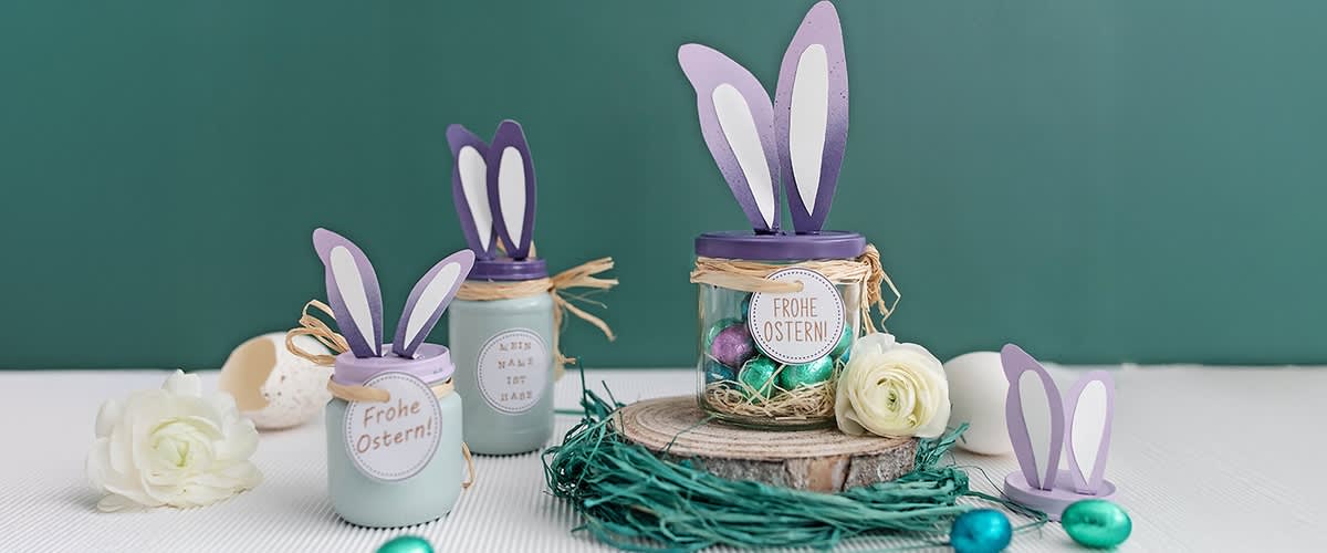 Drei dekorierte Gläser mit lila Hasenohren und der Aufschrift „Frohe Ostern“ stehen auf einem Tisch, umgeben von Ostereiern und Blumen.