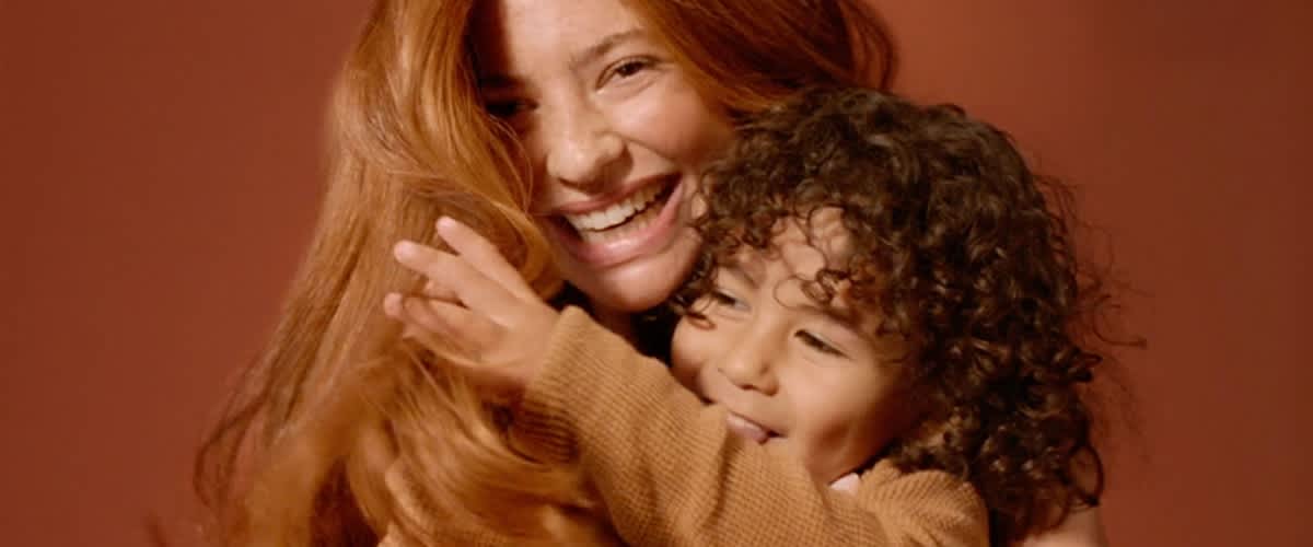 Zwei Personen in warmen Brauntönen, eine Erwachsene hält ein kleines Kind im Arm; logos von L'Oréal Paris oben links und Garnier oben rechts, beide weiße Schrift auf farbigem Hintergrund