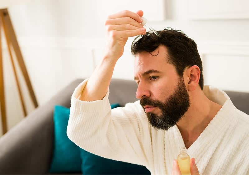 Mann trägt Haaröl auf während er einen Bademantel trägt