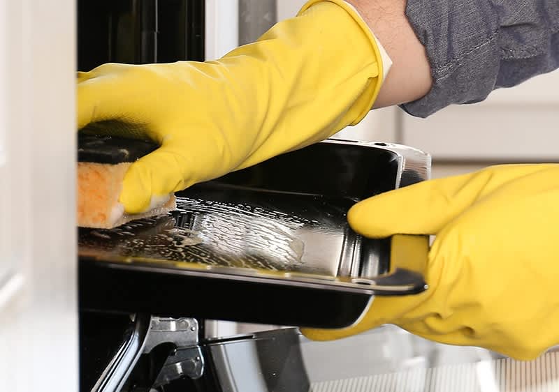 Eine Person mit gelben Gummihandschuhen reinigt ein schwarzes Backblech mit einem Schwamm in einem Ofen.