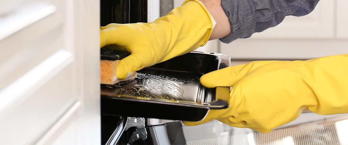 Eine Person mit gelben Gummihandschuhen reinigt ein schwarzes Backblech mit einem Schwamm in einem Ofen.