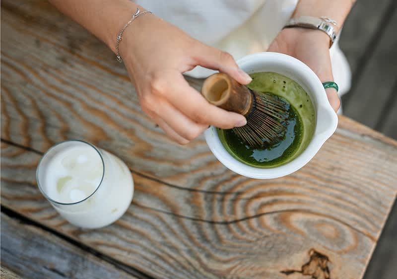 Hände halten eine weiße Schale mit grünem Matcha, ein Bambusbesen wird zum Schlagen des Tees bewegt, daneben ein Glas mit Eis und Milch