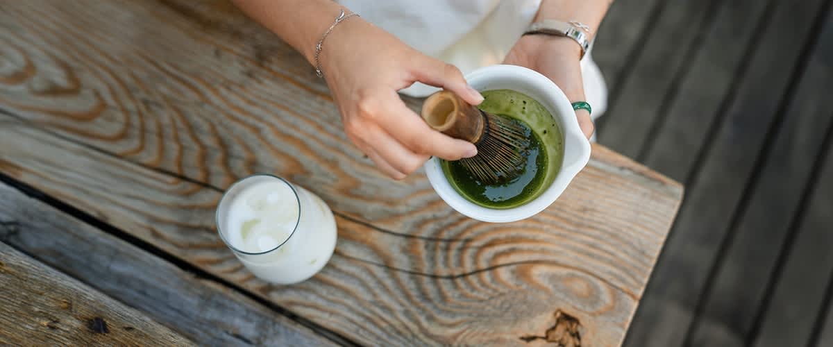 Hände halten eine weiße Schale mit grünem Matcha, ein Bambusbesen wird zum Schlagen des Tees bewegt, daneben ein Glas mit Eis und Milch