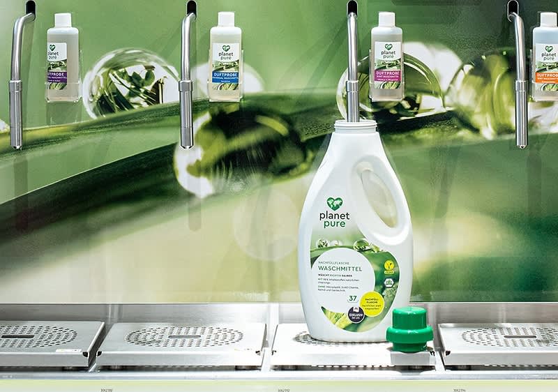 Eine große Flasche Planet Pure Waschmittel steht vor einem grünen Hintergrund mit Pflanzenmotiven. Oben sind vier Spender mit kleinen Flaschen.
