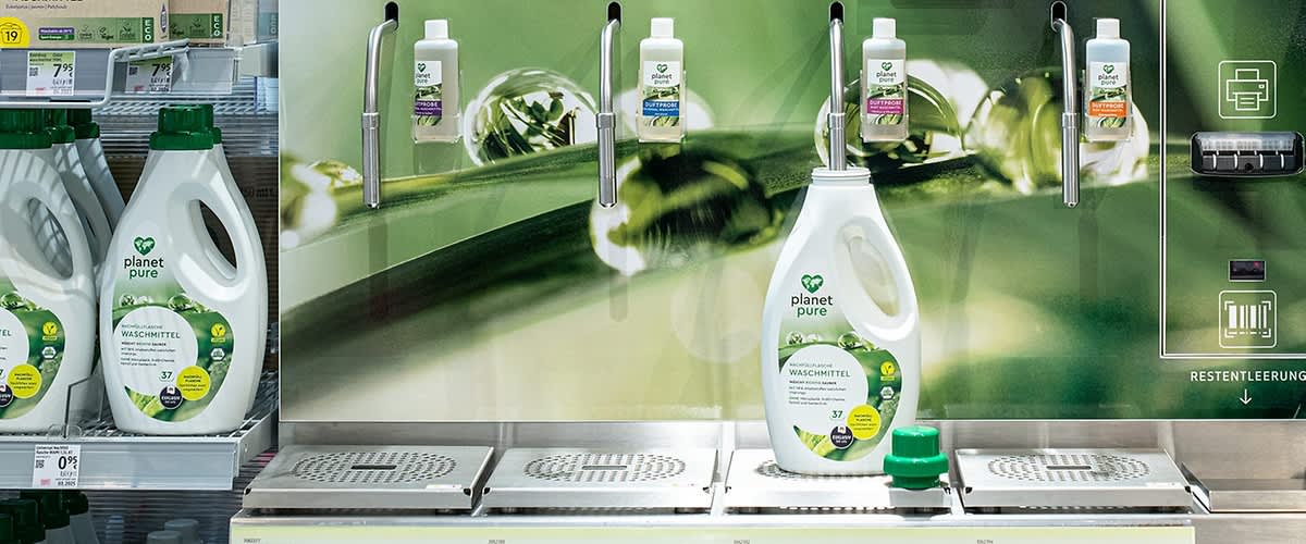 Eine große Flasche Planet Pure Waschmittel steht vor einem grünen Hintergrund mit Pflanzenmotiven. Oben sind vier Spender mit kleinen Flaschen.