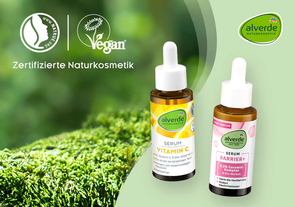 Drei Flaschen Alverde-Serum mit Pipette, jeweils mit unterschiedlicher Aufschrift: Vitamin C, Hyaluron und Barrier. Links oben Logos für zertifizierte Naturkosmetik und vegan. Rechts oben das Alverde-Logo. Im Hintergrund unscharfes Grün.