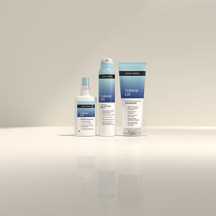 Drei John Frieda Produkte aus der Volume Lift Serie: Ein Spray, ein Schaum und ein Shampoo mit blau-weißer Verpackung und schwarzem Markenlogo