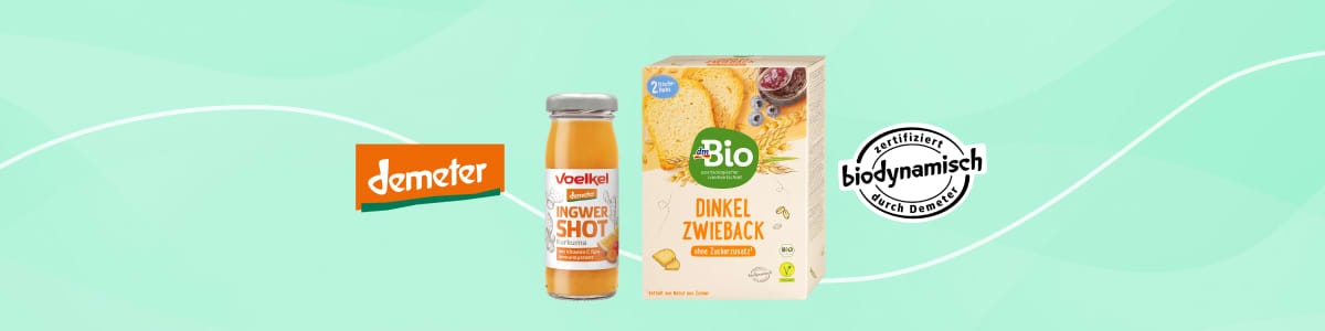 drei Bio-Produkte nebeneinander vor grünem Hintergrund: links oranges Demeter-Logo und eine kleine Glasflasche „Vöslauer Ingwer Shot“, Mitte eine Packung „Bio Dinkel Zwieback“ in gelb-beiger Verpackung, rechts rundes Siegel „biodynamisch“