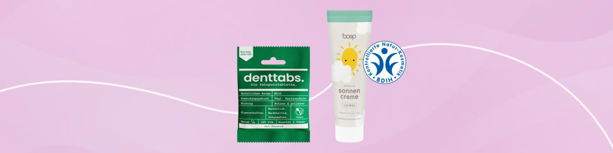 Zwei Produktverpackungen vor hellem Hintergrund: links grüne Packung „denttabs“ mit weißer Aufschrift, rechts weiße Tube mit türkisem Verschluss und Aufschrift „sonnen creme“ sowie Sonnensymbol und blaues Siegel