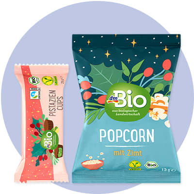 Eine Verpackung von dmBio Popcorn mit Zimt in Blau mit grünen und roten Pflanzenmotiven, daneben eine rosa Verpackung von dmBio Pistazien Cups mit grünen und roten Pflanzenmotiven