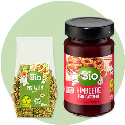 Eine transparente Verpackung mit gehackten Pistazien und der Aufschrift „dmBio Pistazien gehackt“ neben einem Glas mit rotem Inhalt und der Aufschrift „dmBio Himbeere fein passiert“