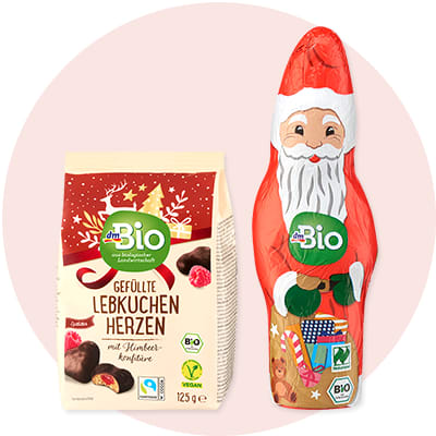 Eine Packung dmBio Gefüllte Lebkuchen Herzen mit Himbeerkonfitüre neben einem Schokoladen-Weihnachtsmann in rotem und weißem Papier mit bunten Geschenken