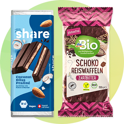 Links eine blaue Verpackung von Share mit der Aufschrift „Coconut Bliss Praliné“, zeigt Schokoladenstücke und Kokosnüsse. Rechts eine Verpackung von dmBio mit der Aufschrift „Schoko Reiswaffeln Zartbitter“, zeigt ein Bio- und Vegan-Siegel.
