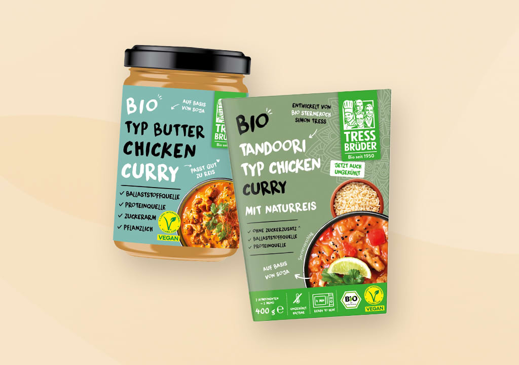 Eine Glasverpackung und eine Kartonverpackung von Tress Brüder Bio-Produkten. Links ein Glas mit Butter Chicken Curry, rechts eine Packung Thai Curry Typ Chicken mit Naturreis. Beide Produkte sind vegan und ohne Zuckerzusatz.