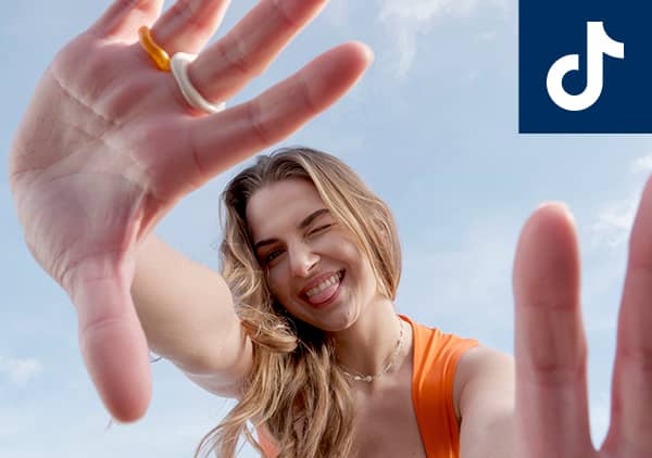 Eine Person in einem orangefarbenen Oberteil streckt ihre Hände mit Ringen in die Kamera, das TikTok-Logo ist oben rechts im Bild
