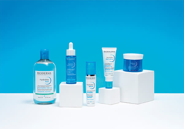 Fünf Bioderma-Produkte auf weißen Podesten vor blauem Hintergrund, darunter eine große Flasche Hydrabio H2O, ein Serum mit Pipette, eine Feuchtigkeitscreme, ein Sonnenschutz und eine blaue Creme in einem Tiegel