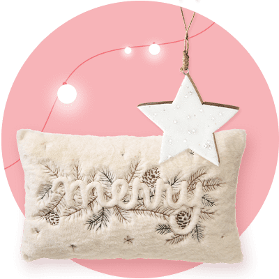 Ein beiges Kissen mit gesticktem Schriftzug „Merry“ und Tannenzweigen, darüber ein weißer Sternanhänger mit goldener Kante vor einem rosa Hintergrund mit Lichterkette